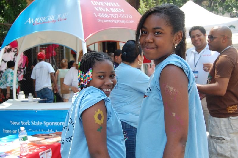 Two mentees in programme t-shirts beside a partner health organisation booth at a street fair.