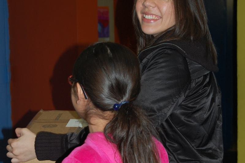 A woman and a girl in a pink coat carrying a cardboard donation box together.