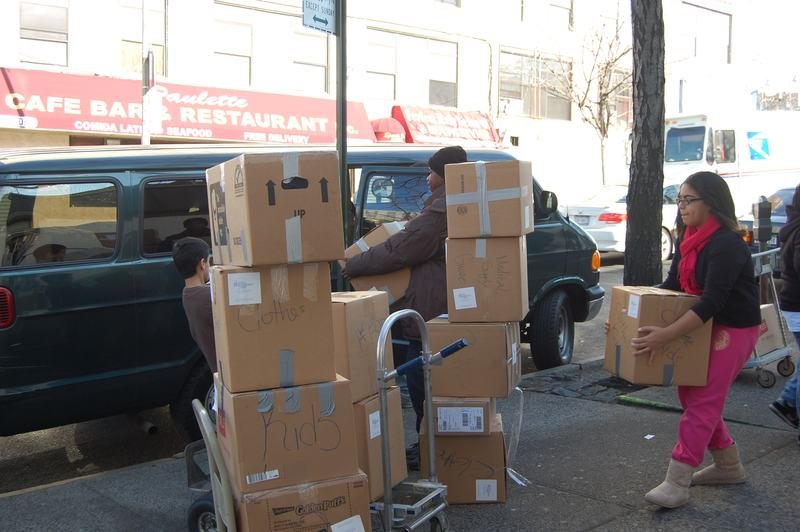 Volunteers wheeling stacked donation boxes along a Bronx street beside a parked van.