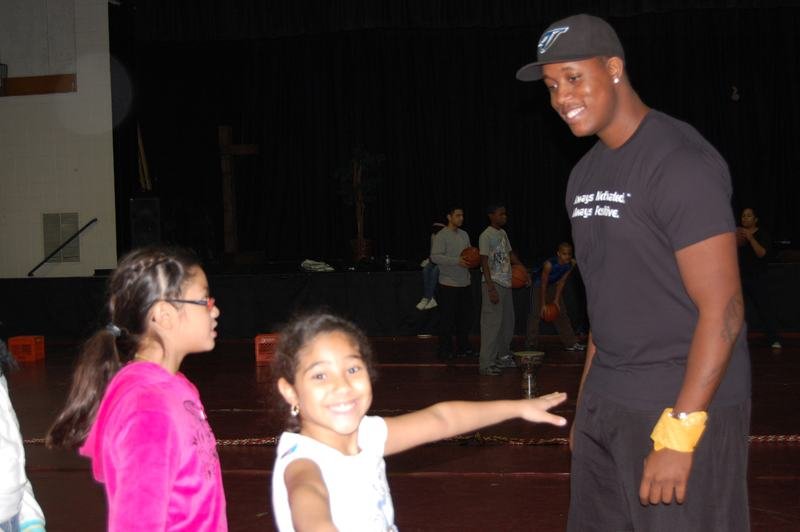A coach in a black cap talks to two young mentees in a dimly lit school gym.