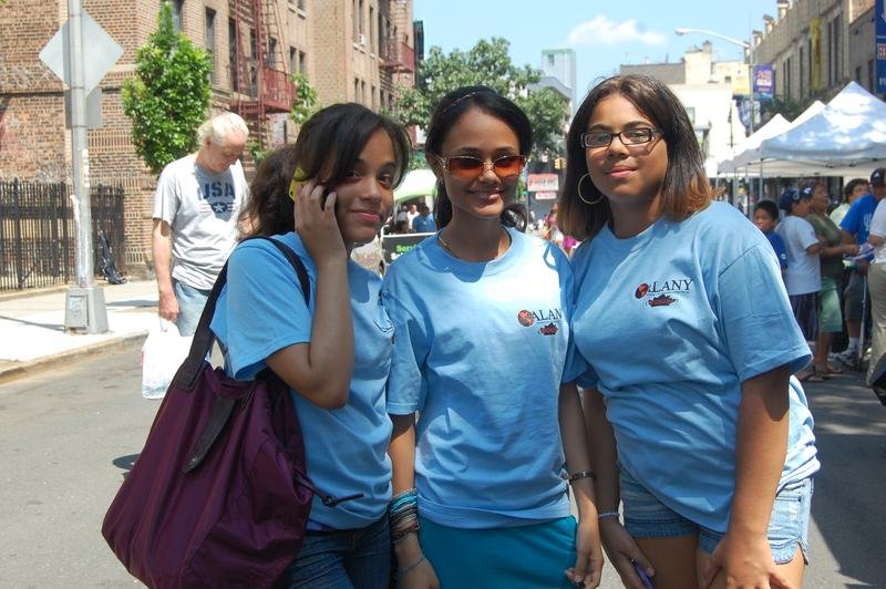 Three ALANY volunteers in blue programme t-shirts on a Bronx street during a fair.