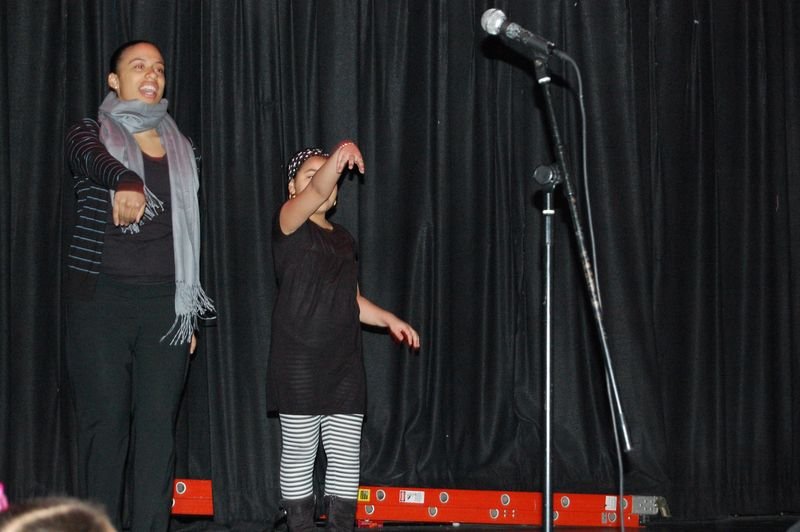 A mentee at a microphone on a dark talent show stage with a staff member beside her.